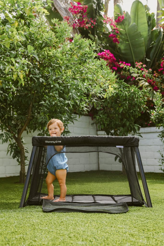 baby playing in travel crib in a backyard with mom sitting nearby photoshoot
