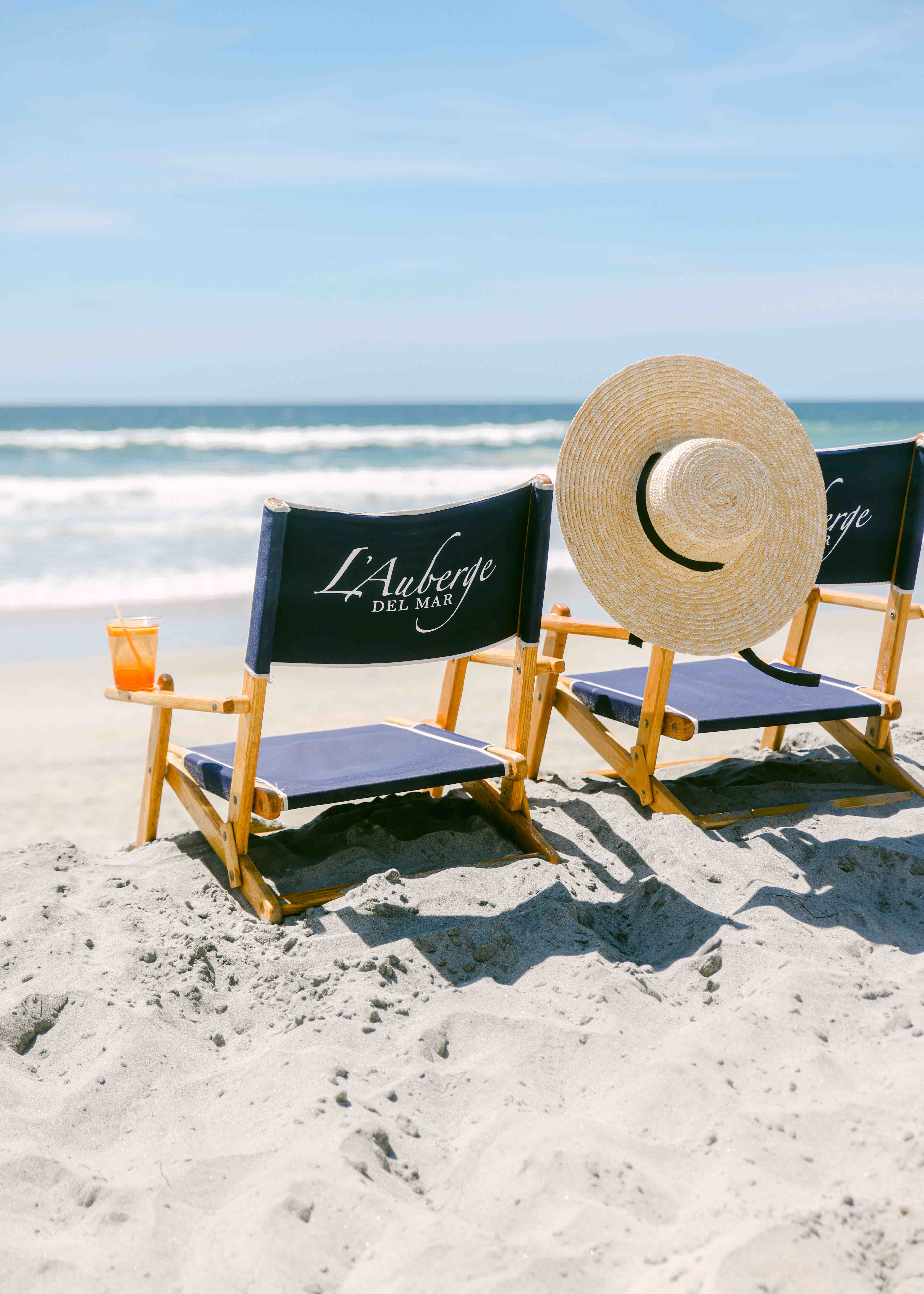Chelsea Loren Southern california photographer for boutique hotel l'auberge del mar with beach chairs on the sand with aperol spritz and large french straw beach hat. Luxury hotel experience photographer.
