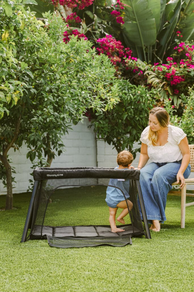 baby playing in travel crib in a backyard with mom sitting nearby photoshoot