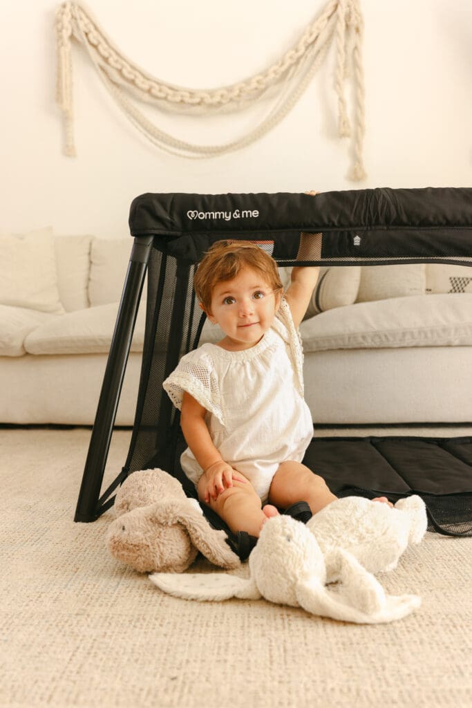 Baby with stuffed animals playing in a lightweight travel crib in a sunlit hotel room natural
