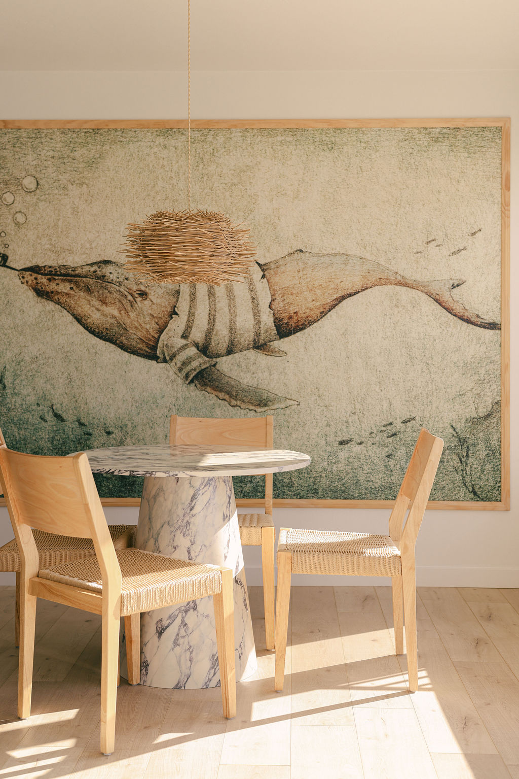 Large framed coastal whale mural above a marble pedestal dining table with wood and rattan chairs at a luxury Airbnb in Carlsbad, California, photographed by Chelsea Loren