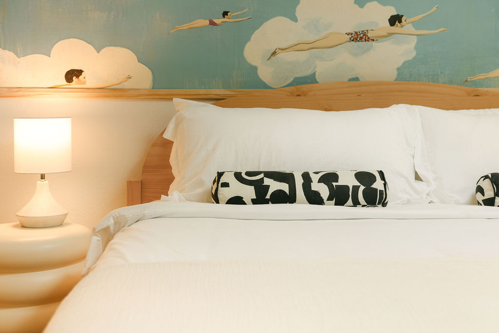 Luxury Airbnb bedroom with swimmer mural headboard, crisp white bedding, and sculptural bedside lamp in Carlsbad, California, photographed by Chelsea Loren