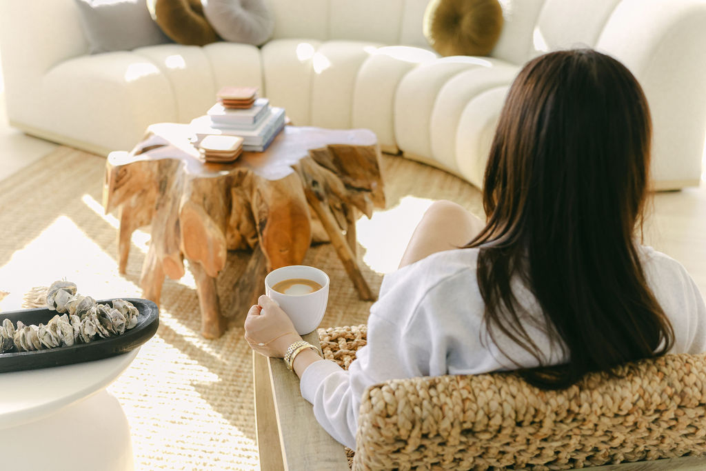 Woman sitting in a woven rattan chair holding a coffee cup beside a curved white sofa and live-edge wood stump table at a luxury Airbnb in Carlsbad, California, photographed by Chelsea Loren