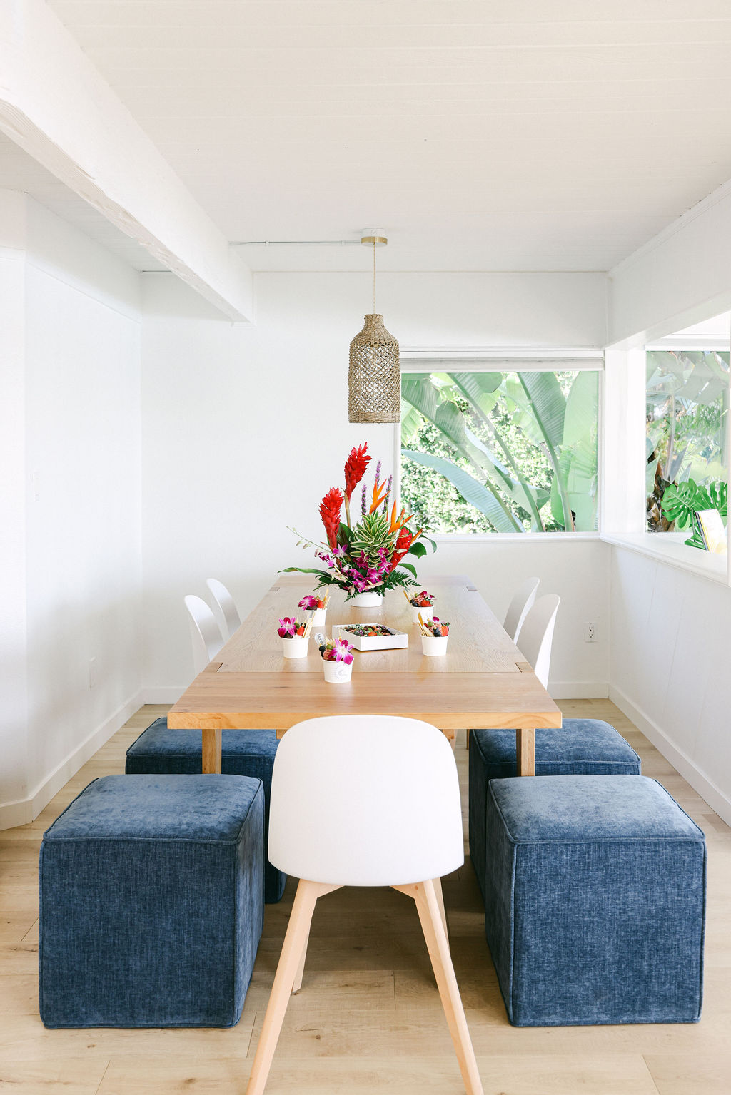 Bright open-plan dining room with rattan pendant light, tropical flower centerpiece, and blue velvet ottomans at a luxury Carlsbad Airbnb, photographed by Chelsea Loren