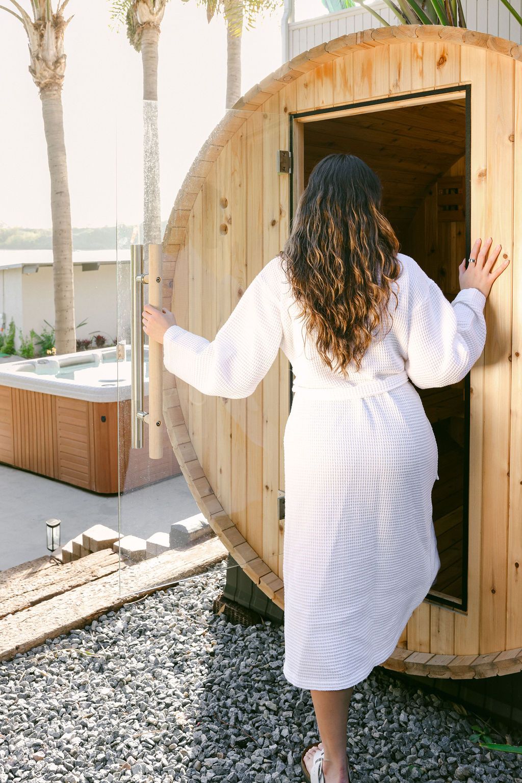 Woman in a white waffle robe stepping into an outdoor barrel sauna with a hot tub and palm trees at a luxury Carlsbad Airbnb, photographed by Chelsea Loren