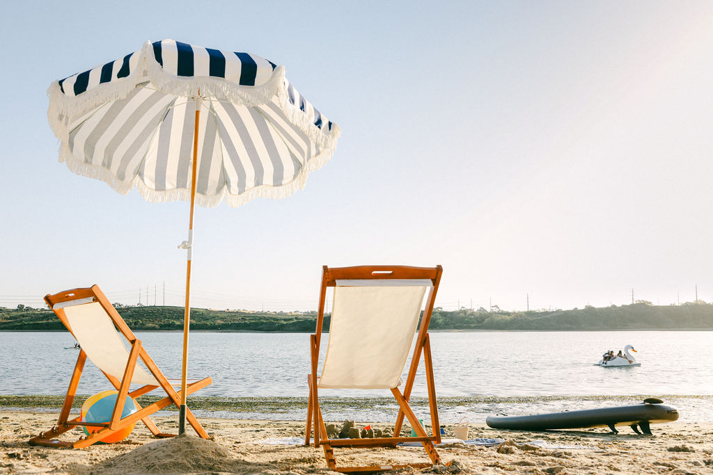Beachfront Airbnb amenity with wooden deck chairs and striped umbrella on the sandy shore with a paddleboard and swan paddleboat, photographed by Chelsea Loren