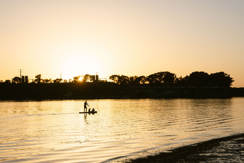 Silhouette of paddle boarders at golden hour on the Carlsbad lagoon, dreamy sunset lifestyle photography for a luxury waterfront short-term rental by Chelsea Loren