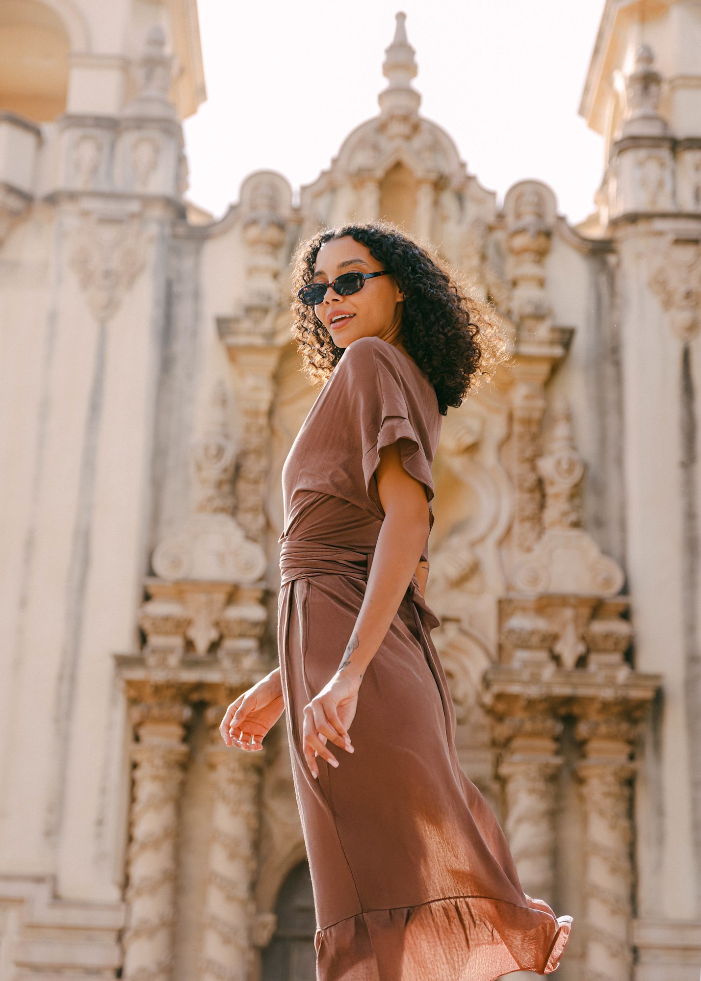 Fashion lookbook model in brown wrap dress mid-stride against ornate California architecture | Chelsea Loren Creative Director
