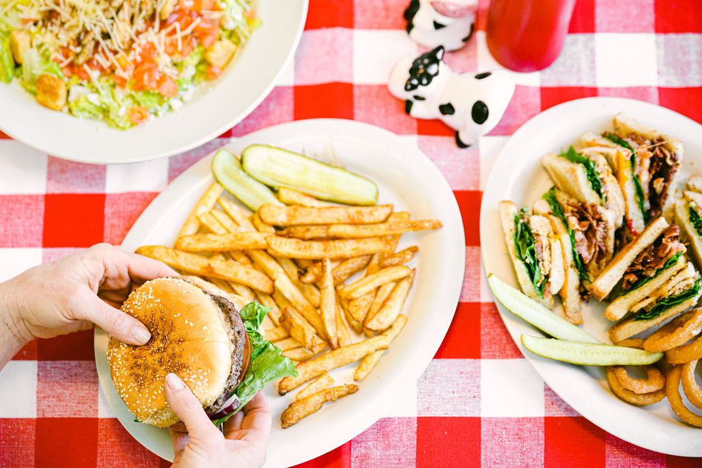 Top-down flatlay of burger, fries, club sandwich, salad, and cow salt and pepper shakers on red checkered tablecloth at MooMoo's Burger Barn Lodi