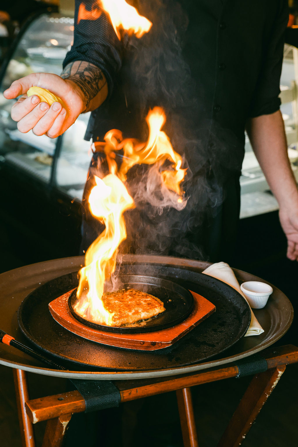 Flaming saganaki being prepared tableside with dramatic orange flames and chef's tattooed hands at Papapavlo's Bistro in Lodi California