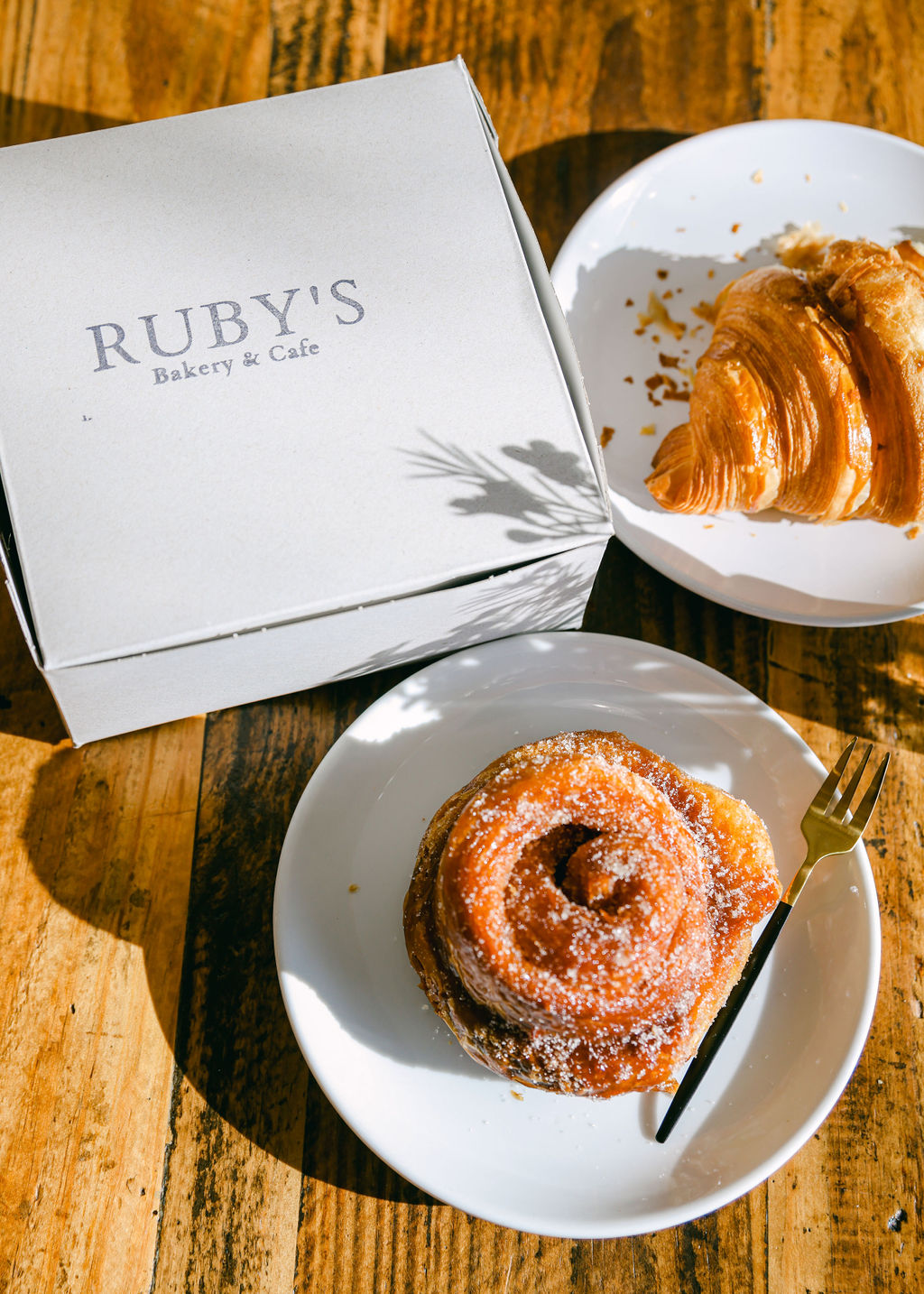 Ruby's Bakery pastry box with sugar bun and croissant in dappled sunlight with gold fork on marble