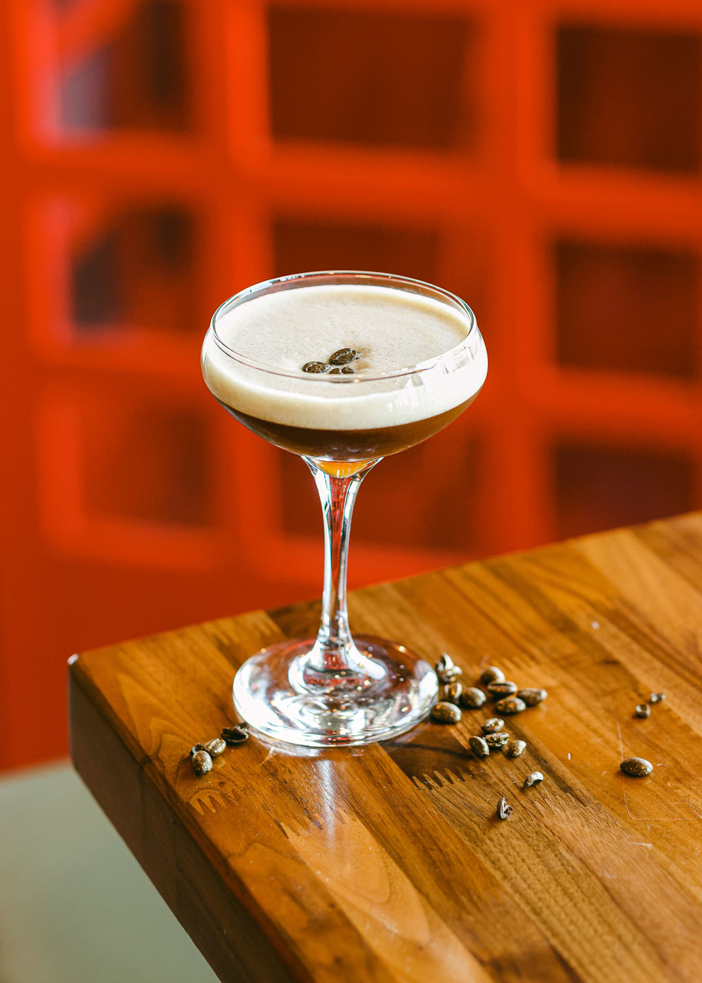 Espresso martini in coupe glass perched on wood ledge with scattered coffee beans and red backdrop at The Oxford Kitchen Lodi