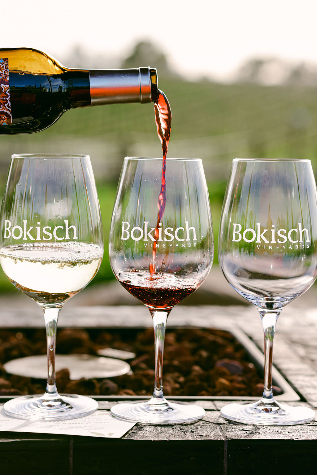 Three Bokisch Vineyards wine glasses on outdoor surface with red wine being poured into center glass and vineyard backdrop