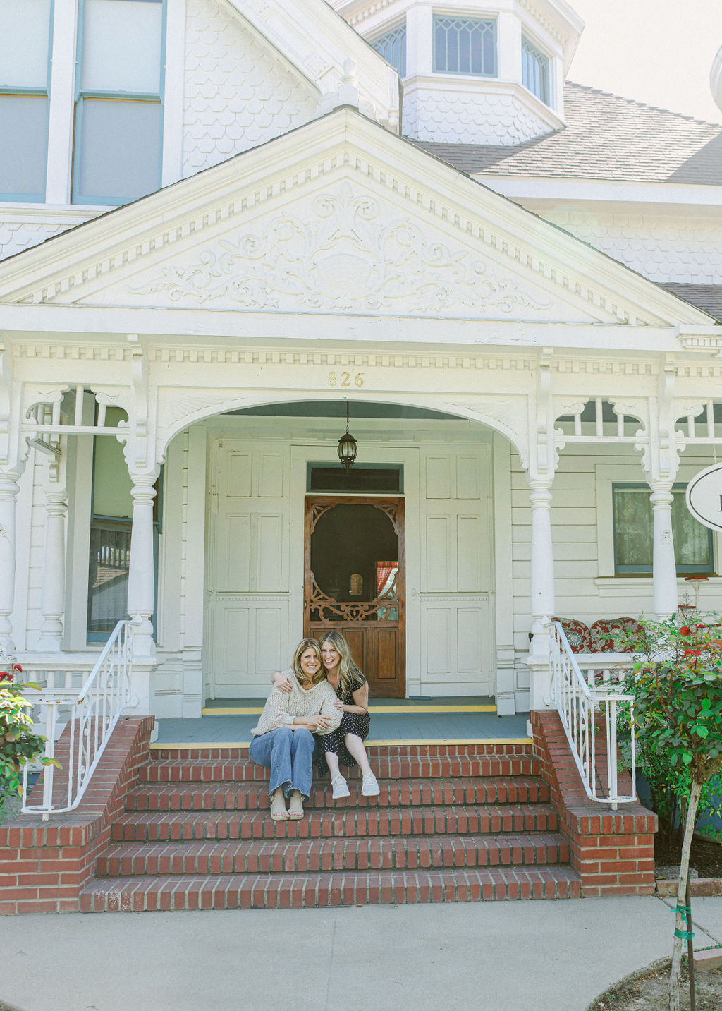 Two women sitting together on brick steps of the ornate white Victorian Lodi Hill House Inn facade