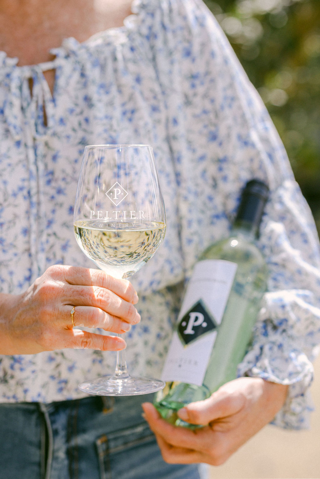 Woman in blue floral blouse holding Peltier Winery glass of white wine and bottle in sun-dappled outdoor setting Lodi